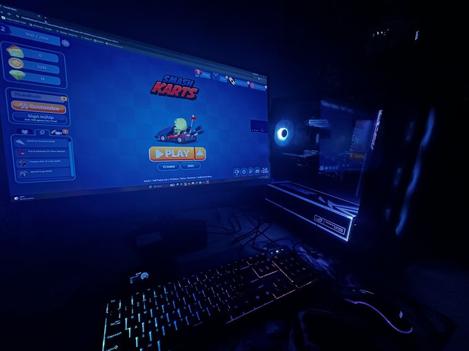 A dark gaming setup features a curved monitor displaying the title screen for 'Crash Karts' above a backlit keyboard and a gaming PC case with blue lighting.