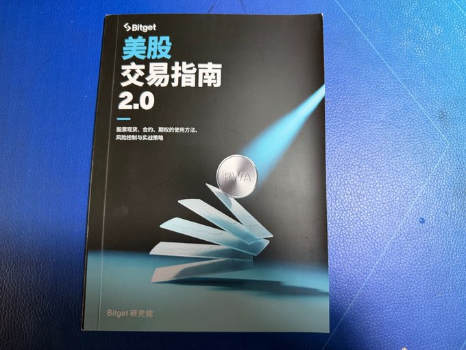 A Bitget research guide book titled '美股交易指南 2.0' (US Stock Trading Guide 2.0) in Chinese, with an RWA coin graphic on the cover, photographed on a blue surface.