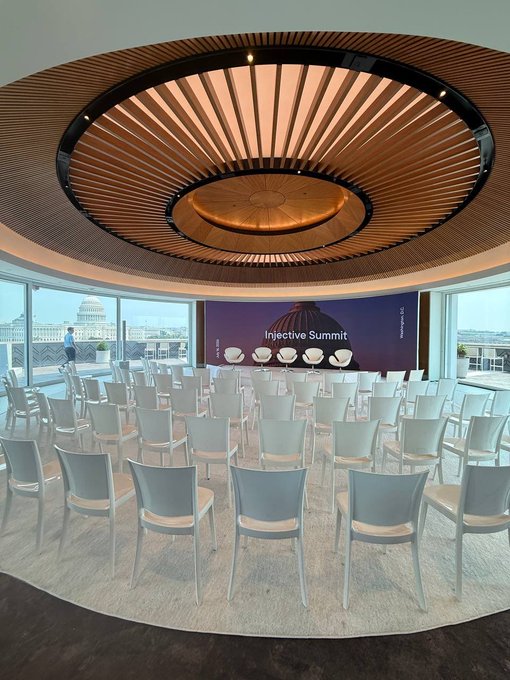 A modern circular conference room with rows of chairs facing a screen displaying 'Injective Summit', featuring a striking wooden slat ceiling design.
