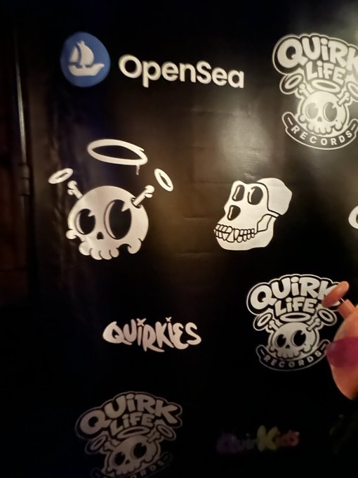 A black wall displays promotional logos including "OpenSea," "Quirk Life Records," and "Quirkies," alongside a Bored Ape-style skull character with halos, while a person's hand is visible on the right side, capturing the NFT community gathering mentioned in the post.