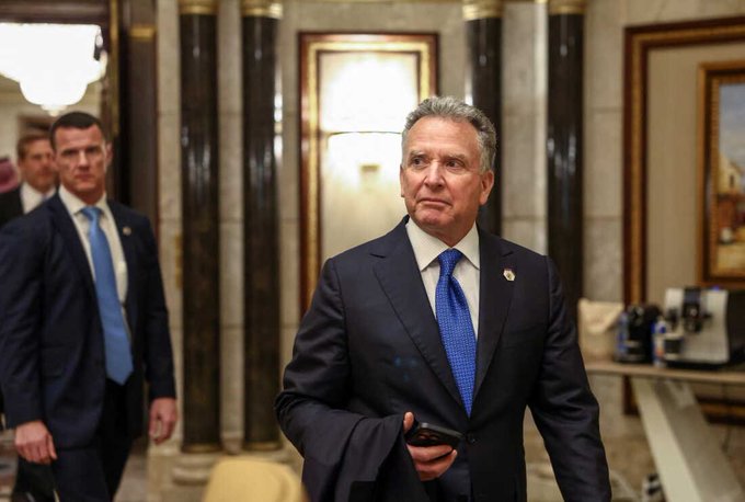 Steve Witkoff, Trump's special envoy, walks through an ornate marble hallway while carrying a phone, with another man in a suit following behind him. The image accompanies news of Witkoff's disclosure of $107 million in 2025 income from a holding company tied to World Liberty Financial, a crypto venture in which he previously held a stake.