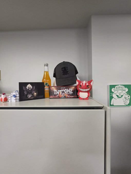 A photo of a white shelf displaying various collectibles including a Mythical (MYTH) orange soda bottle, a black cap, a 'GRINDC' green box, an 'INTU' figurine, and other small toys.