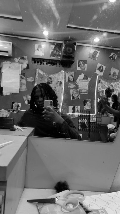 A person with dreadlocks takes a mirror selfie with their phone in what appears to be a salon, surrounded by hair style posters and images pinned to the walls. A pair of scissors and papers rest on the counter below, suggesting a haircut appointment—fitting the "Dread Head" caption highlighting their hairstyle.