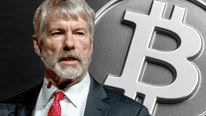 An older bearded man in a suit and red tie stands in front of a large Bitcoin logo, appearing to speak or give a statement.