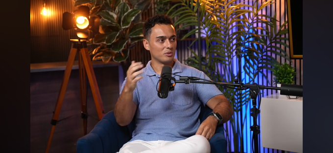 A man in a light blue polo shirt sits in a podcast studio, speaking into a professional microphone with a warm spotlight and tropical plants in the background, aligning with the post's mention of doing an in-person podcast.