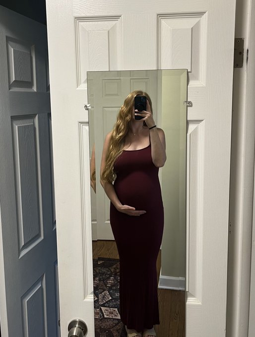 A pregnant woman with long red hair in a burgundy maxi dress takes a mirror selfie while holding her belly, standing in front of a white door.