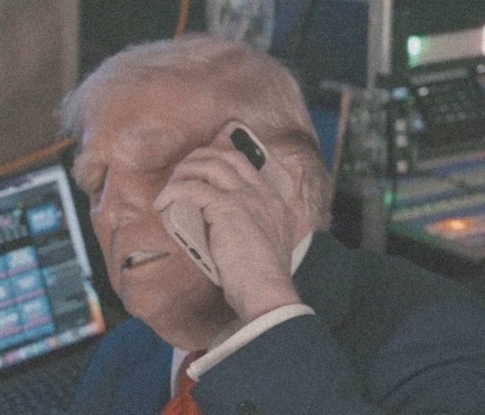 The image shows what appears to be a trader on the floor of a stock exchange, smiling while speaking on a mobile phone amid monitors displaying market data. This candid moment of a finance professional working the trading floor visually reinforces the stock market enthusiasm conveyed by the caption "Pump it son," suggesting bullish market activity and confident trading.