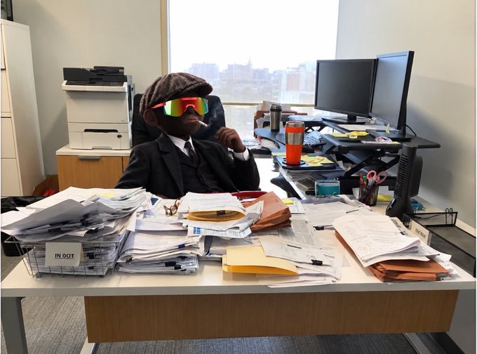 A man in a suit wearing a flat cap, reflective ski goggles, and smoking a cigarette sits at an office desk overflowing with stacks of papers and documents.