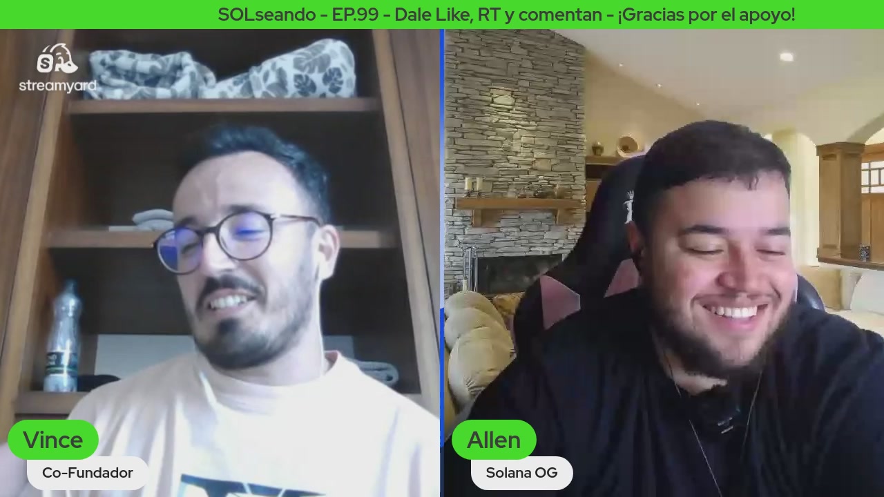 The image shows two men sitting next to each other, both smiling at something or someone off-camera. They are positioned close together, with one man's face being slightly more visible than the other. The background appears to be a living room setting, featuring a couch and a chair. There is also an object in the scene that has the word "solana" written on it.