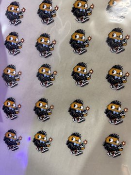 A sheet of stickers featuring a small, cartoonish pixel-art character with a black hood, large orange eyes, and what appears to be a weapon or tool, repeated in a grid pattern.