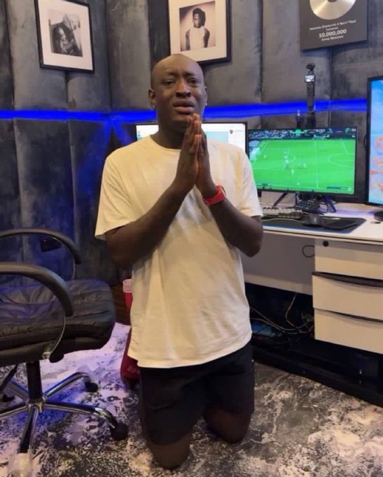 A man in a white t-shirt kneels on one knee with his hands clasped together in prayer, in a room with framed photos, a TV showing a soccer match, and blue LED lighting.
