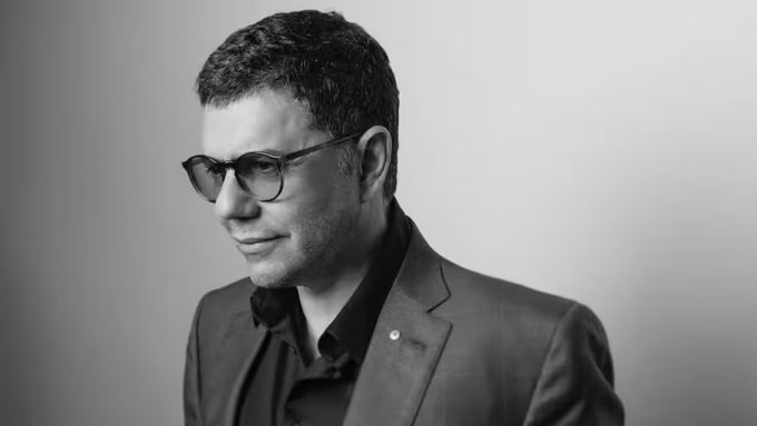 A black and white portrait of a man with short dark hair wearing glasses, a dark shirt, and a suit jacket, looking off to the side against a plain background.