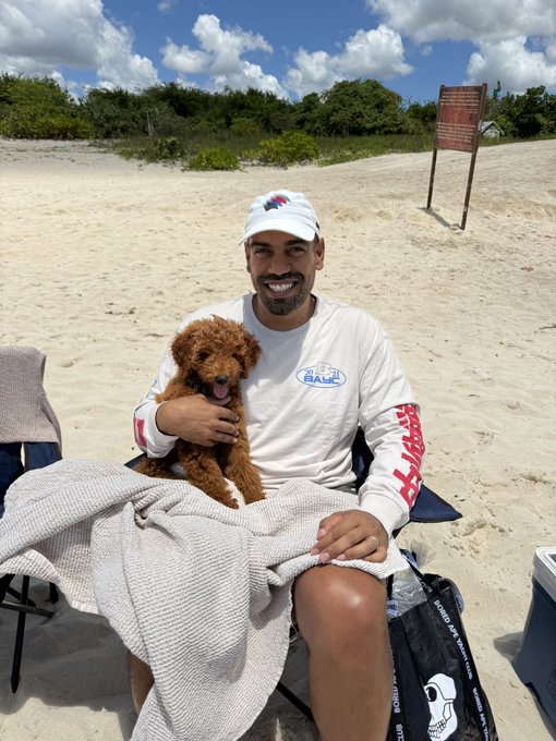 A smiling man wearing a white cap and long-sleeve shirt sits on a sandy beach holding a brown poodle, with a towel draped over his lap.