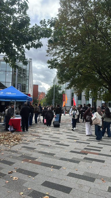 An outdoor plaza scene shows people gathered near a blue tent labeled 'Tillery' on a paved walkway, with modern glass buildings and trees in the background.