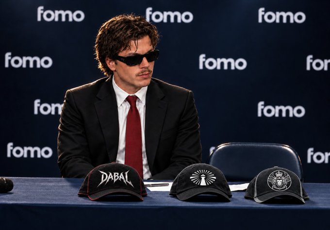 A man in a suit, tie, and sunglasses sits at a table with a "fomo" backdrop, with three baseball caps displayed in front of him.