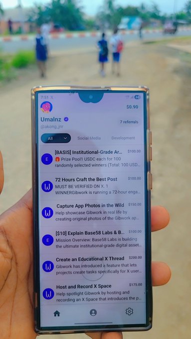 The image shows a person holding up their cell phone, which displays several messages from different people. The messages include one from @BybitOfficial asking for help creating a thread or contest, as well as another message from @gib_work asking for assistance with a task on the app.gib.work/. The person's hand is visible in front of them, and they are standing next to a road where several people can be seen walking by.