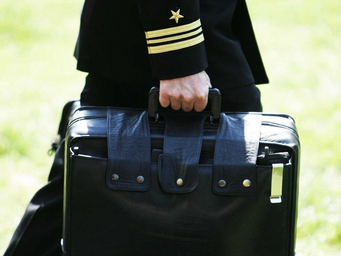 A close-up of a military officer's hand gripping a black leather briefcase, with the officer's uniform sleeve displaying gold stripes and a star insignia.