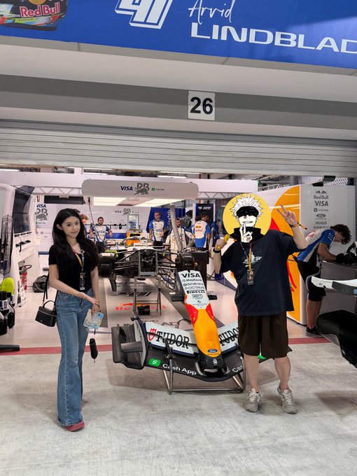 Two people pose in front of a Visa Cash App RB Formula 1 car display in a garage area marked '26', with sponsor branding for Red Bull, HUGO, and Arvid Lindblad visible.