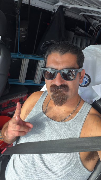 A man with a dark goatee, wearing sunglasses and a gray tank top, gestures with his hand while sitting inside what appears to be a vehicle or workshop.