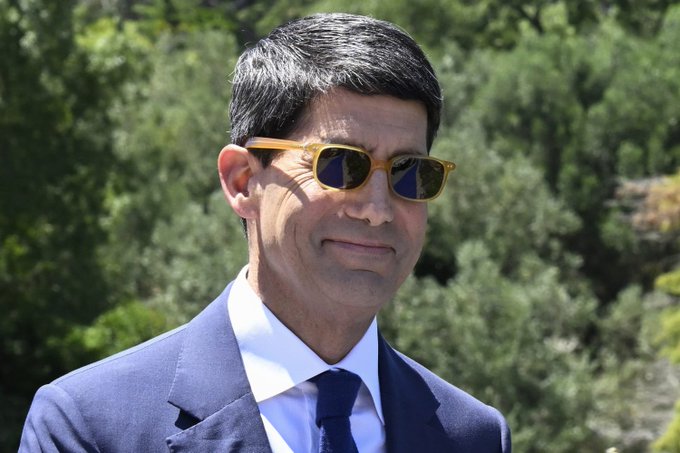 A man in a dark suit, white shirt, and sunglasses with yellow-tinted frames smiles subtly against a backdrop of green foliage, likely outdoors. The image likely accompanies the post as a visual of a person reacting to the news, possibly representing Fed Chair Kevin Warsh, whose comments on easing inflation risks are mentioned as a catalyst for the cryptocurrency rally.