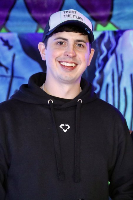 A smiling young man wearing a black hoodie and a black-and-white cap reading "TRUST THE PLAN" stands in front of a colorful graffiti wall, posing as a proud member of the Bored Ape Yacht Club community.
