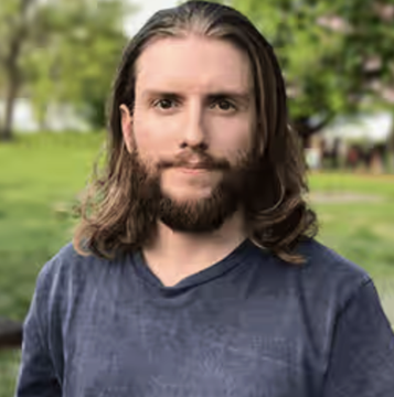 A man with long dark hair and a full beard, wearing a gray t-shirt, stands in what appears to be a park setting, likely Chris Barrett from Chainlink discussing how regulatory delays on crypto rules disadvantage the US by benefiting its competitors.