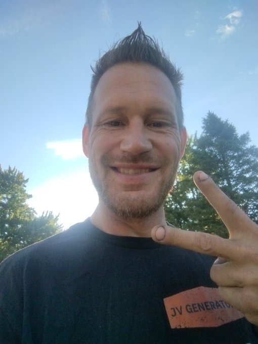 A smiling man with a short beard and spiked hair takes a selfie outdoors against a clear blue sky, flashing a peace sign with his fingers while wearing a black shirt featuring an orange "JV GENERAL" logo patch. The sunny, upbeat "GM in the AM" vibe is perfectly captured in his cheerful expression and bright morning lighting.