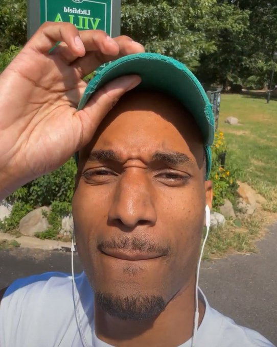 A young man wearing a green cap and white earphones takes a close-up selfie outdoors, with trees and a street visible in the background.