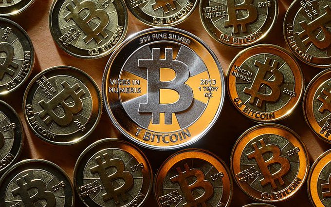 A close-up photograph of multiple silver and gold Bitcoin commemorative coins, with the central coin inscribed '.999 FINE SILVER', 'VIRES IN NUMERIS', '2013 1 TROY OZ', and '1 BITCOIN'.