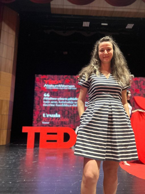 A young woman with long wavy hair wearing a black and white striped dress stands on a stage at a TEDxAtakumWomen event, with a screen displaying 'Ursula Ede A. Gain' behind her.