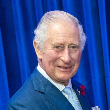 King Charles III, dressed in a dark suit with a red poppy pin, smiles warmly against a royal blue backdrop during an event where he convened AI industry executives to discuss the existential risks of artificial intelligence and advocate for international oversight.