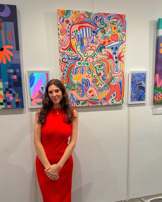 A woman in a sleeveless red dress standing in an art gallery in front of a large, colorful abstract painting featuring faces and organic shapes, with additional artwork displayed on adjacent walls.