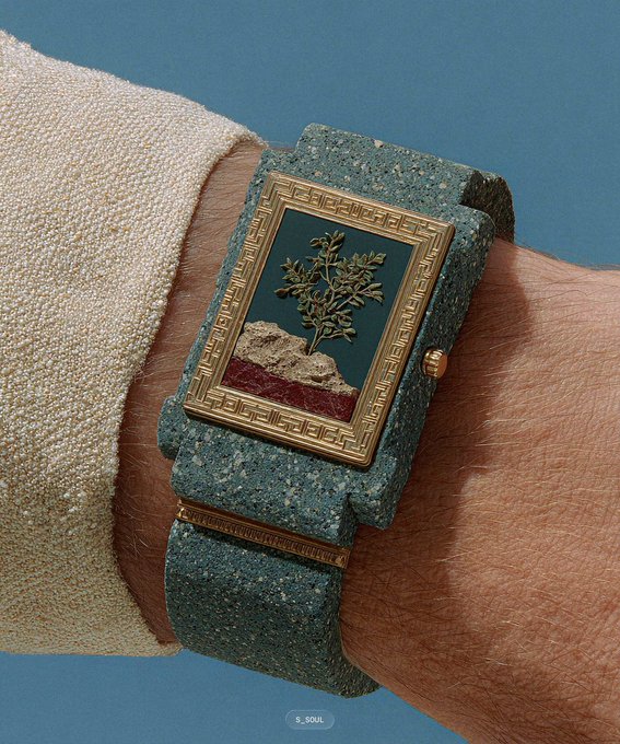 A wrist sports a distinctive square watch with a gold Greek-key frame, dark green dial featuring a tree and landscape motif, mounted on a textured green stone-like strap.