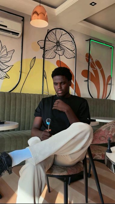 A young man in a black t-shirt and beige pants sits on a chair in a stylish café, holding what appears to be a hookah pipe, with decorative floral wall art behind him.