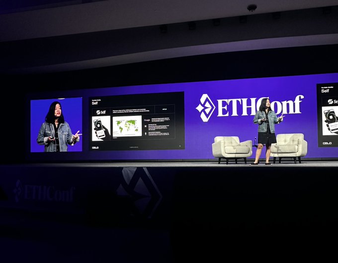 The image shows a woman standing behind a podium at an event, giving a presentation or speech. The stage has a large screen displaying information related to EthConf, which is a conference focused on identity solutions. The speaker's name tag reads "Elizabeth Peng", and she appears to be discussing the company's growth as a leading identity solutions provider. 

There are two chairs visible in the image, one located near the front of the stage and another further back. A person can also be seen standing behind the podium, possibly assisting with the presentation or taking notes.