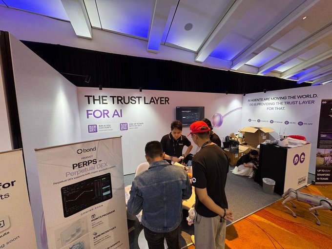 An event booth displays signage reading "THE TRUST LAYER FOR AI" and "Qbond PERPS Perpetual DEX", with several people gathered around a counter in a well-lit conference hall.