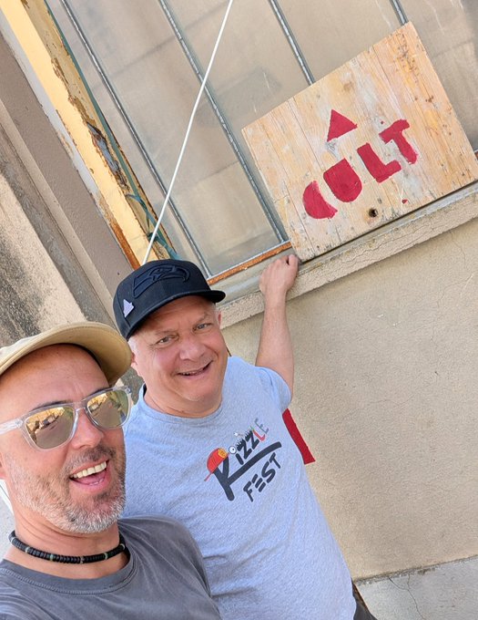 The image features two men standing next to each other, both wearing baseball caps. They are posing for a photo with smiles on their faces. The background of the picture includes a building that has a sign hanging from its window, which reads "Cult CryptoArt". This suggests that they might be at an event or gathering related to cryptocurrency and art.