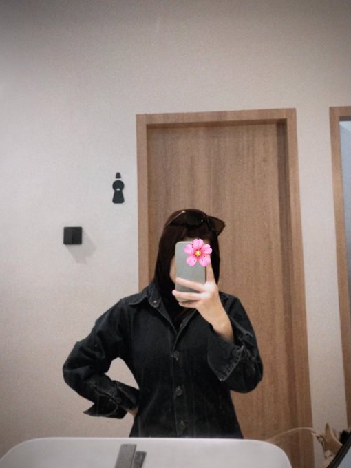 A mirror selfie of a person wearing a black denim jacket and sunglasses on their head, taking the photo with a phone that has a pink flower case in front of a wooden door.