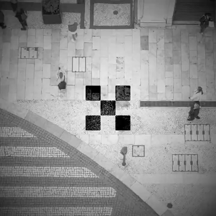 This black-and-white aerial photograph captures a bird's-eye view of a pedestrian plaza featuring Portuguese-style cobblestone paving, where several people can be seen walking or standing near what appear to be metal grates and utility covers. The image bears no visible text, numbers, or logos, and there is no discernible connection to "OKX" within the scene itself.