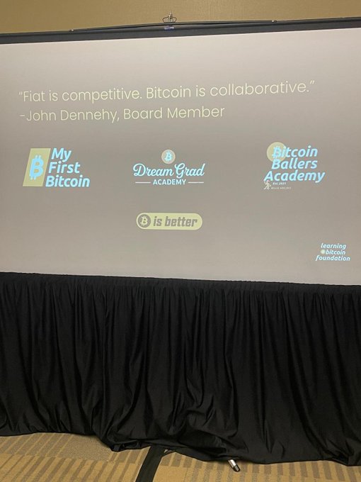 A projection screen at a Bitcoin-related event displays the quote 'Fiat is competitive. Bitcoin is collaborative.' by John Dennehy, Board Member, alongside logos for My First Bitcoin, Dream Grad Academy, and Bitcoin Ballers Academy, sitting above a black draped table.