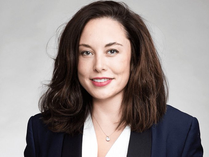 A professional portrait of a young woman with shoulder-length brown hair, wearing a navy blazer over a white blouse and a small pendant necklace, smiling against a gray background.