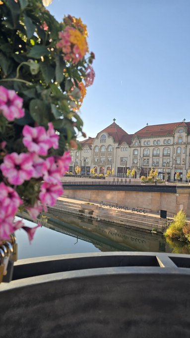 A scenic view of a historic European building with a red roof across a calm canal/river, framed by pink flowers and hydrangeas in the foreground under a clear blue sky.