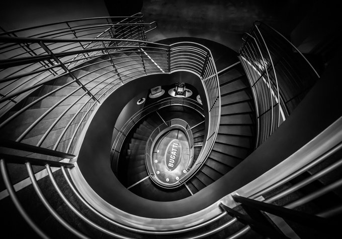 Black and white photograph looking down a spiral staircase with curved railings, featuring a Bugatti emblem at the bottom center.