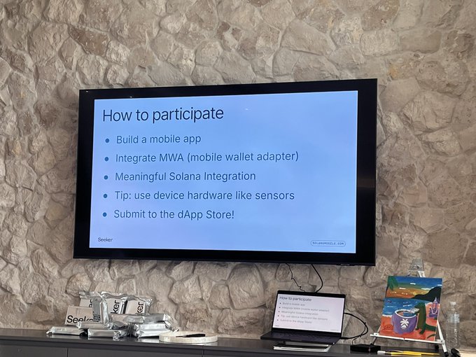 The image shows a television screen displaying information about participating in the @solanamobile Week, which is an event that encourages people to use their smartphones for social good projects. The screen provides instructions on how to participate by building a mobile app or integrating Solana into existing applications. It also suggests tips and tricks for using device hardware like sensors to enhance the user experience. Additionally, there are books and a laptop nearby, which could be used as resources during the event.