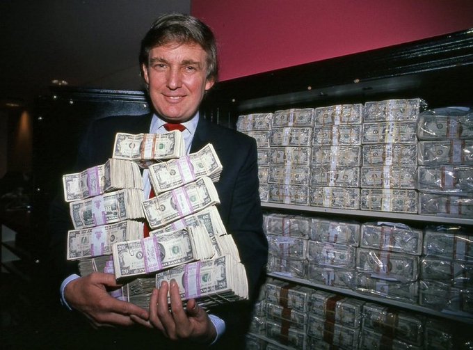 A photograph of a smiling man in a suit holding stacks of US dollar bills, with massive stacks of bundled cash filling shelves behind him.