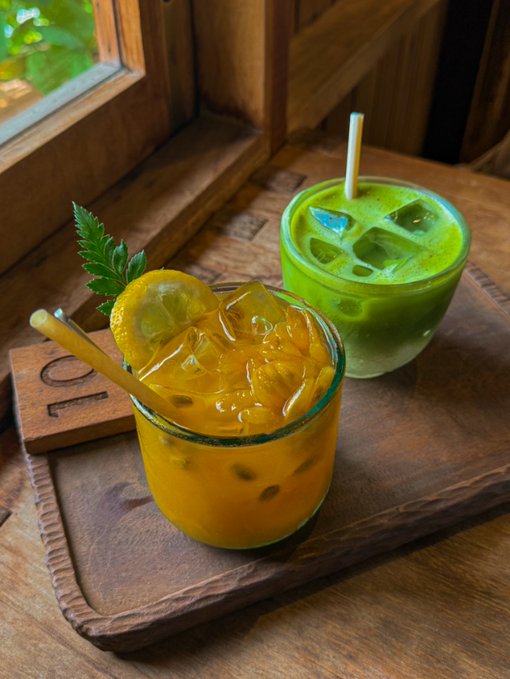 Two iced beverages—one yellow/orange and one green—served in glasses with paper straws on a wooden tray beside a window, garnished with a lemon slice and mint leaf.