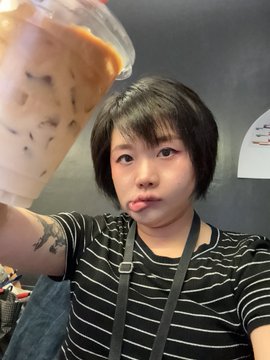 A young woman with short black hair, wearing a striped shirt and lanyard, holds up a clear plastic cup containing a milky iced beverage (appears to be Thai milk tea).