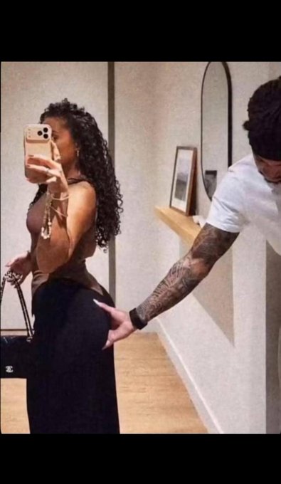 A mirror selfie of a woman with long curly hair wearing a brown top and black skirt, holding a phone. A man with tattooed arms and a white t-shirt stands beside her, reaching toward her.