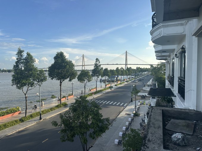 A scenic waterfront street lined with trees runs alongside a wide river, with a large cable-stayed bridge visible in the distance under a partly cloudy sky, while a white multi-story building with balconies stands on the right.