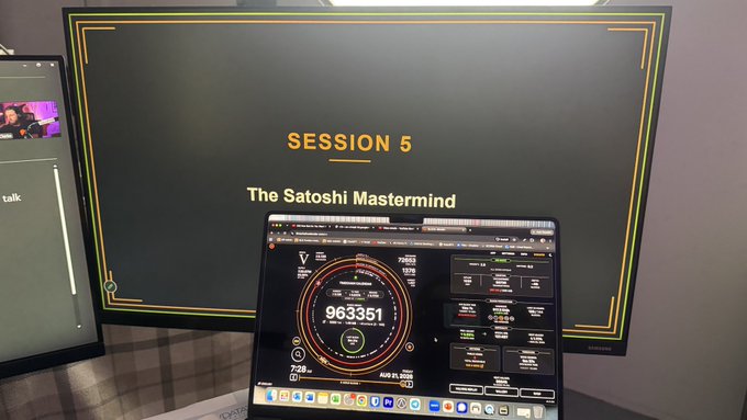 Photograph of a laptop displaying what looks like a node monitoring dashboard with a block height of 963351, placed in front of a large monitor showing "SESSION 5 - The Satoshi Mastermind" in an event setting.